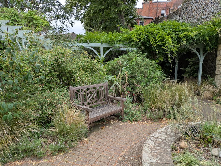 A small, lush garden corner featuring a weathered wooden bench with a decorative backrest situated on a curved brick pathway. Surrounding the bench are dense, overgrown plants, including shrubs with s